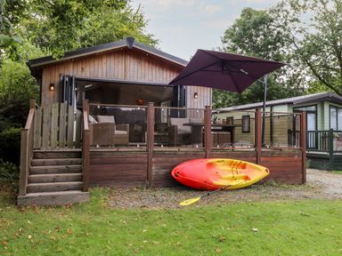 A wooden lodge with outdoor seating and a purple umbrella next to a red and yellow kayak at Loubi's Lakeside Lodge M1 Fallbarrow Park near Bowness-On-Windermere