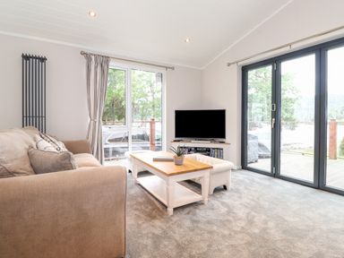 A living room with a beige sofa a white coffee table and a television near large windows at Loubi's Lakeside Lodge M1 Fallbarrow Park near Bowness-On-Windermere