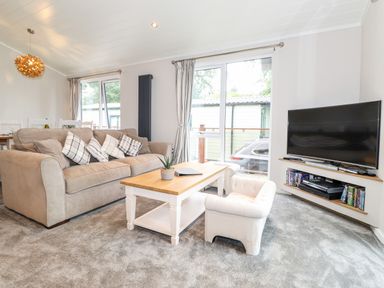 A living room with a beige sofa cushions a white coffee table an armchair a TV on a corner stand and dining table with chairs at Loubi's Lakeside Lodge M1 Fallbarrow Park near Bowness-On-Windermere
