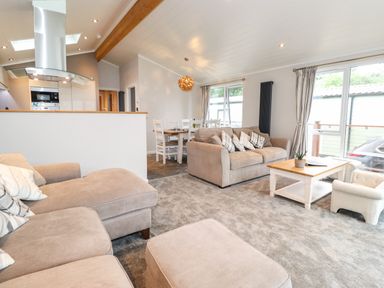 A living room with beige sofas a wooden coffee table white dining chairs and a kitchen area at Loubi's Lakeside Lodge M1 Fallbarrow Park near Bowness-On-Windermere