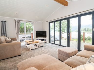 A living room with sofas a coffee table a TV and large windows overlooking a deck and water at Loubi's Lakeside Lodge M1 Fallbarrow Park near Bowness-On-Windermere