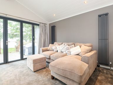 A living room with a beige sectional sofa and matching ottoman near glass sliding doors at Loubi's Lakeside Lodge in M1 Fallbarrow Park near Bowness-On-Windermere
