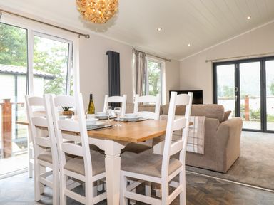 A dining area with a wooden table and white chairs next to a living room with sofas and large windows at Loubi's Lakeside Lodge in M1 Fallbarrow Park near Bowness-On-Windermere