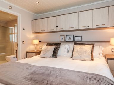 A bedroom with a large bed pillows bedside tables lamps and overhead cabinets with a bathroom visible through an open door at Loubi's Lakeside Lodge M1 Fallbarrow Park near Bowness-On-Windermere