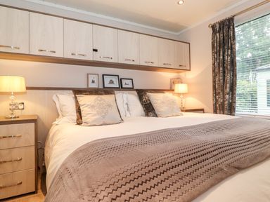 A bedroom with a large bed two bedside tables with lamps wall cabinets and a window with curtains at Loubi's Lakeside Lodge M1 Fallbarrow Park near Bowness-On-Windermere
