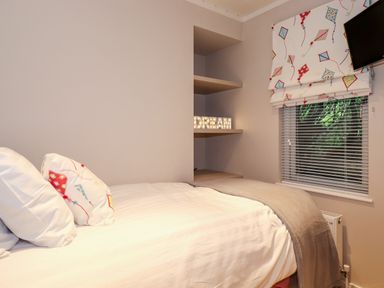A bedroom with a bed pillows and a blanket near shelves with a DREAM light and a window with blinds and a patterned roman shade at Loubi's Lakeside Lodge M1 Fallbarrow Park near Bowness-On-Windermere