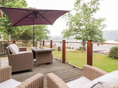 An outdoor deck with a wicker sofa and chairs a table with glasses and bottles under a large umbrella overlooking a lakeside park at Loubi's Lakeside Lodge M1 Fallbarrow Park near Bowness-On-Windermere