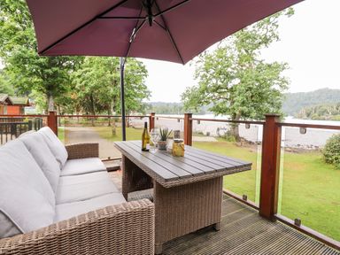 An outdoor seating area with a wicker sofa and table under a large umbrella next to a lake at Loubi's Lakeside Lodge M1 Fallbarrow Park near Bowness-On-Windermere