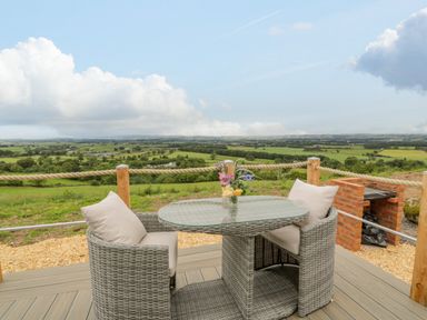 An outdoor patio with a glass top table and two cushioned chairs a small vase with flowers on the table a brick barbecue and a countryside view at Laird Lodge 2 in Lochmaben