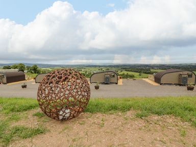 An outdoor view with a large metal spherical sculpture on grass and modern pod cabins in the background at Laird Lodge 2 in Lochmaben