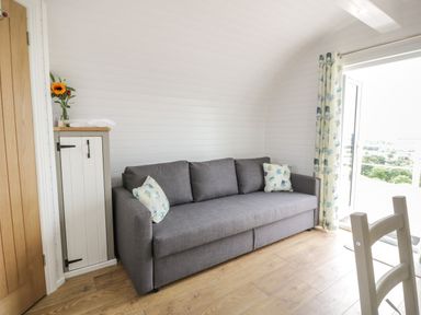 A small living area with a gray sofa and floral cushions next to a wooden cabinet and glass door with floral curtains at Laird Lodge 3 in Lochmaben