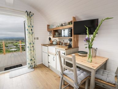 A small kitchen area with a wooden table and chairs next to a sliding door with floral curtains opening to a balcony at Laird Lodge 3 in Lochmaben