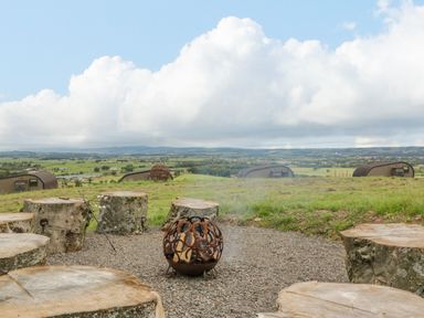 A circle of tree stump seats around a fire pit with a rural landscape and cabins in the background at Laird Lodge 3 in Lochmaben