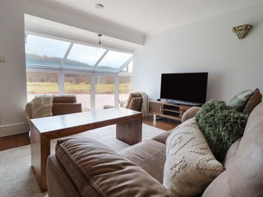 A living room with a sofa, armchairs, wooden coffee table, television, and large windows at Tan Yr Allt in Dolanog near Meifod Powys