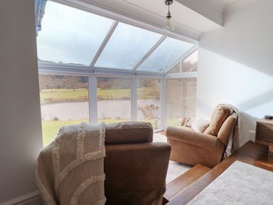 A sunroom with two leather armchairs facing windows overlooking a lake and grassy landscape at Tan Yr Allt in Dolanog near Meifod Powys