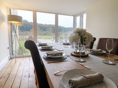 A dining room with a wooden table set with plates napkins glasses and a flower centerpiece by large windows at Tan Yr Allt in Dolanog near Meifod Powys
