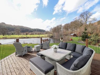 An outdoor deck with wicker furniture including chairs and a table overlooking a lake and trees at Tan Yr Allt in Dolanog near Meifod Powys