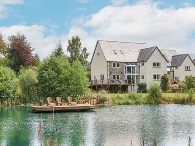 A house with a wooden deck and outdoor chairs by a pond surrounded by trees at Tan Yr Allt in Dolanog near Meifod Powys