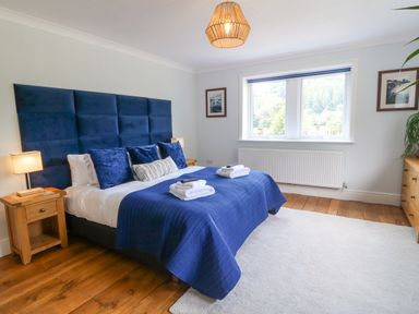 A bedroom with a blue bedspread and pillows wooden bedside tables lamps a wooden dresser and a large window at Tan Yr Allt in Dolanog near Meifod Powys