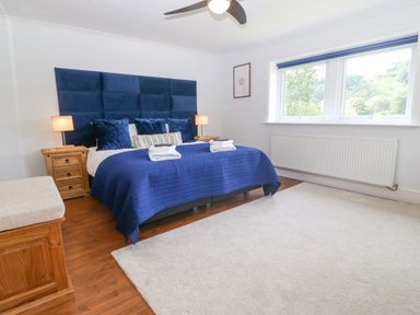 A bedroom with a king size bed with blue bedding and pillows two wooden nightstands with lamps a wooden bench a white radiator and a large window at Tan Yr Allt in Dolanog near Meifod Powys