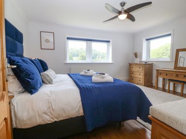 A bedroom with a bed with blue bedding two windows a wooden dresser and dressing table at Tan Yr Allt in Dolanog near Meifod Powys