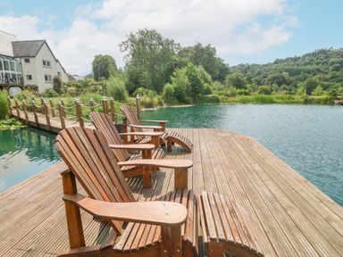 A wooden deck with chairs overlooking a lake and wooden bridge at Tan Yr Allt in Dolanog near Meifod Powys