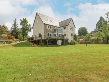 An exterior view of a two-story house with a wooden deck and large lawn surrounded by trees at Tan Yr Allt in Dolanog near Meifod Powys