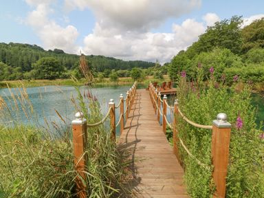 A wooden walkway with rope railings leading to a deck with chairs by a pond at Tan Yr Allt in Dolanog near Meifod Powys