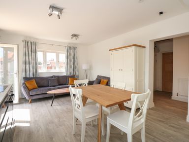 A living room with a wooden dining table with four white chairs gray sofas with orange cushions a white cupboard and a window with curtains at Jolly Sailors Retreat - Wells next the Sea in Wells-Next-The-Sea