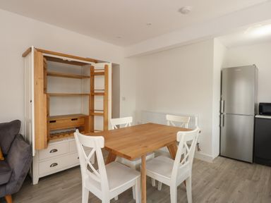 A dining area with a wooden table and four white chairs next to an open wooden cabinet and a silver refrigerator at Jolly Sailors Retreat - Wells next the Sea in Wells-Next-The-Sea
