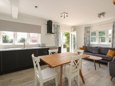 A kitchen and dining area with a wooden table and white chairs next to a gray sofa and windows at Jolly Sailors Retreat - Wells next the Sea in Wells-Next-The-Sea
