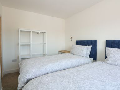 A bedroom with two twin beds with blue headboards and white patterned bedding with a nightstand and white shelving unit in Jolly Sailors Retreat in Wells-Next-The-Sea