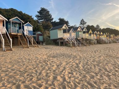 A row of small beach huts elevated on stilts with wooden staircases on a sandy beach with trees in the background at Jolly Sailors Retreat - Wells next the Sea in Wells-Next-The-Sea