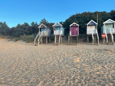 A row of beach huts on stilts with stairs leading to the doors on a sandy beach with trees in the background at Jolly Sailors Retreat - Wells next the Sea