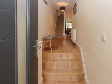 A hallway with wooden stairs leading to a carpeted staircase a bench on the left wall a radiator on the right wall and a dining area visible at the end at Mimosa in Dawlish