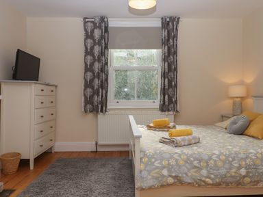 A bedroom with a bed with grey and yellow bedding a chest of drawers with a television a window with patterned curtains and a lamp on a bedside table at Mimosa in Dawlish