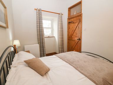 A bedroom with a bed white sheets beige pillow and blanket a wooden door window with curtains and a bedside lamp at Ty Gwenyn in Rhydwyn