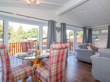 A dining area with a glass table and plaid chairs next to a living room with gray sofas and curtains at Fell Foot Lodge in Fellside 9 Limefitt Park near Troutbeck