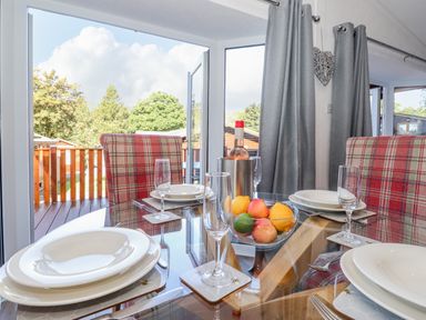 A dining table set with white plates glasses a bowl of fruit and a bottle of wine next to plaid chairs and glass doors at Fell Foot Lodge in Fellside 9 Limefitt Park near Troutbeck
