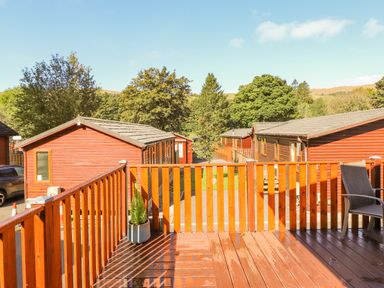 A wooden deck with a potted plant and chair overlooking red cabins and trees at Fell Foot Lodge Fellside 9 Limefitt Park near Troutbeck