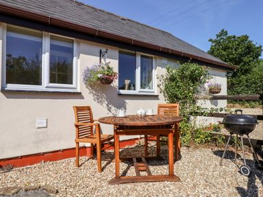 A patio area with a round wooden table and two wooden chairs next to a house with two windows and hanging flower baskets at Little Rowan at Rowan Park Farm in Ashwater