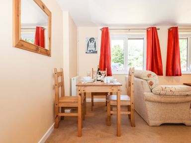 A dining area with a wooden table set for four and beige sofa near windows with red curtains at Little Rowan at Rowan Park Farm in Ashwater