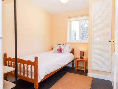 A small bedroom with a single wooden bed a floral pillow a wooden bedside table with a lamp and a window with a view of a stone wall at Little Rowan at Rowan Park Farm in Ashwater