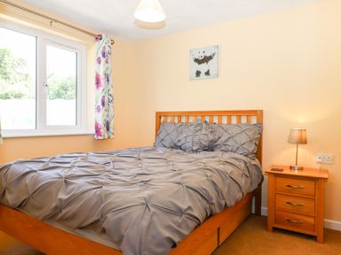 A bedroom with a wooden bed and nightstand with a lamp and a window with floral curtains at Little Rowan at Rowan Park Farm in Ashwater