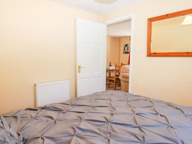 A bedroom with a bed covered in a gray bedspread a white door open to a room with a dining table and chairs and a wooden framed mirror on the wall at Little Rowan at Rowan Park Farm in Ashwater