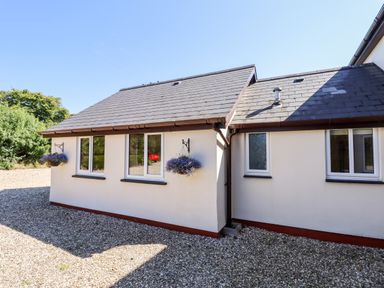 The exterior of a house with white walls multiple windows and hanging flower baskets on a gravel area at Little Rowan at Rowan Park Farm in Ashwater