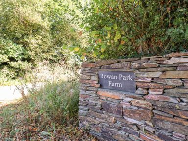A stone wall with a Rowan Park Farm sign surrounded by grass and trees at Little Rowan at Rowan Park Farm in Ashwater