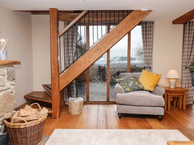A living room with a gray armchair with pillows near wooden stairs with a basket of logs and a side table lamp at Little Orchard in Ashburton Down near Ashburton