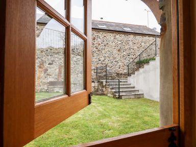 A view through an open wooden window showing a grassy courtyard with stone steps and a stone building at Little Orchard in Ashburton Down near Ashburton