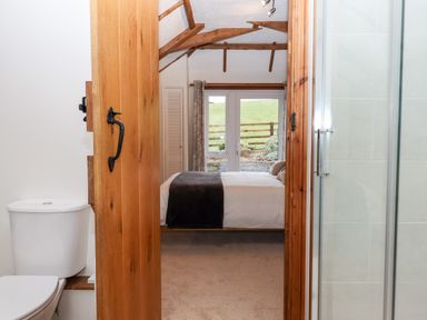 A bedroom with a bed and windows showing a green field visible through an open door leading from a bathroom with a toilet and shower at Little Orchard in Ashburton Down near Ashburton